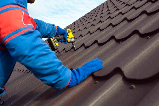 Man worker uses a power drill to attach a cap metal roofing job with screws.