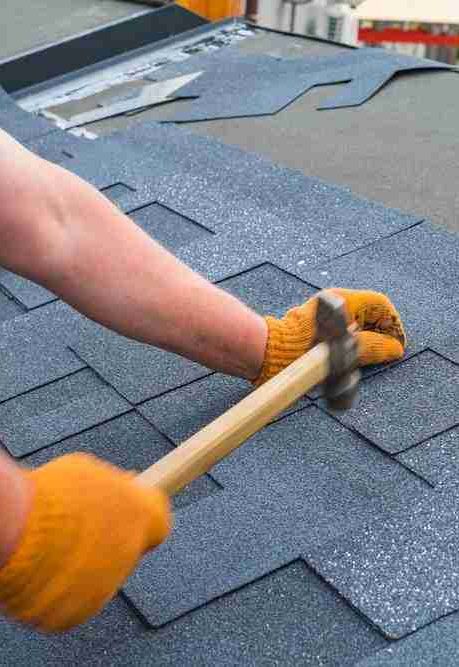 Workers hands installing bitumen roof shingles using hammer in nails.